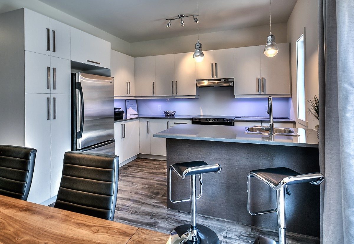 Cuisine d’une maison du Quartier Eaux-Vives : armoires blanches, réfrigérateur en acier, îlot avec tabourets noirs
